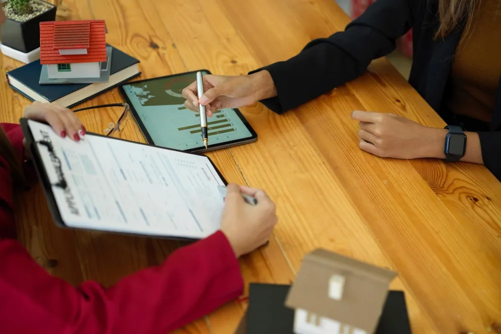 Hands reviewing a property form and tablet beside a small house model (illustrative)